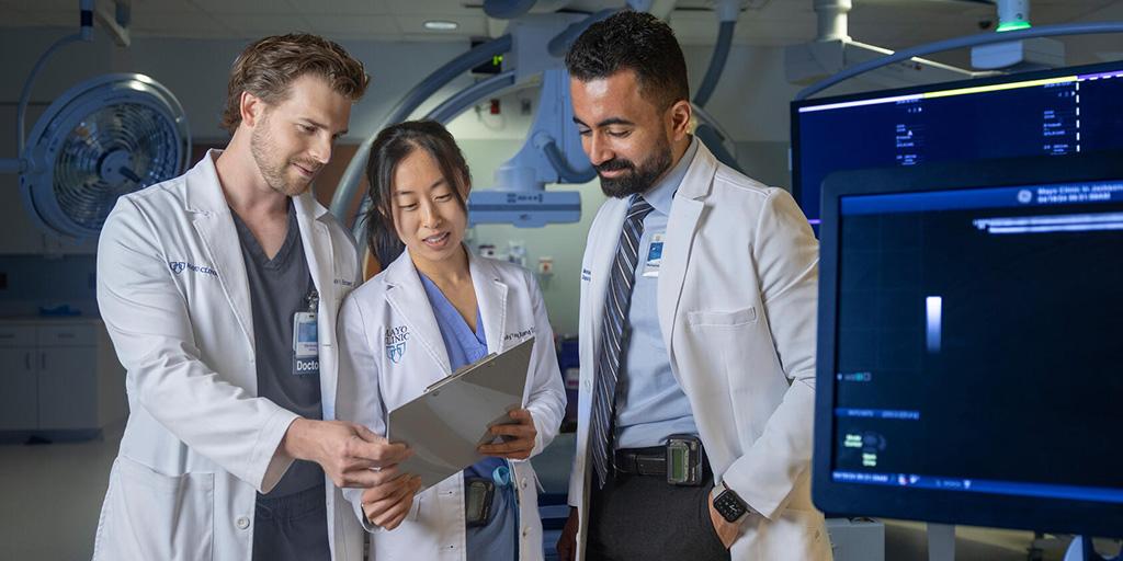 Three radiology residents, wearing white coats, look at a clipboard together while standing in a procedure room.