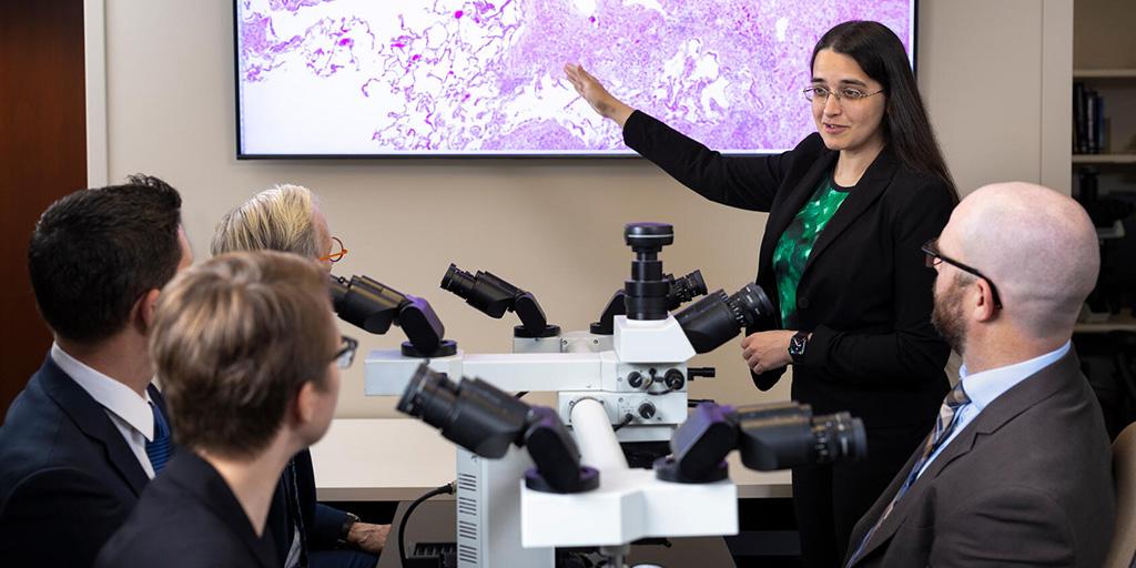 A doctor is pointing to a screen on the wall during a presentation in front of four people seated.