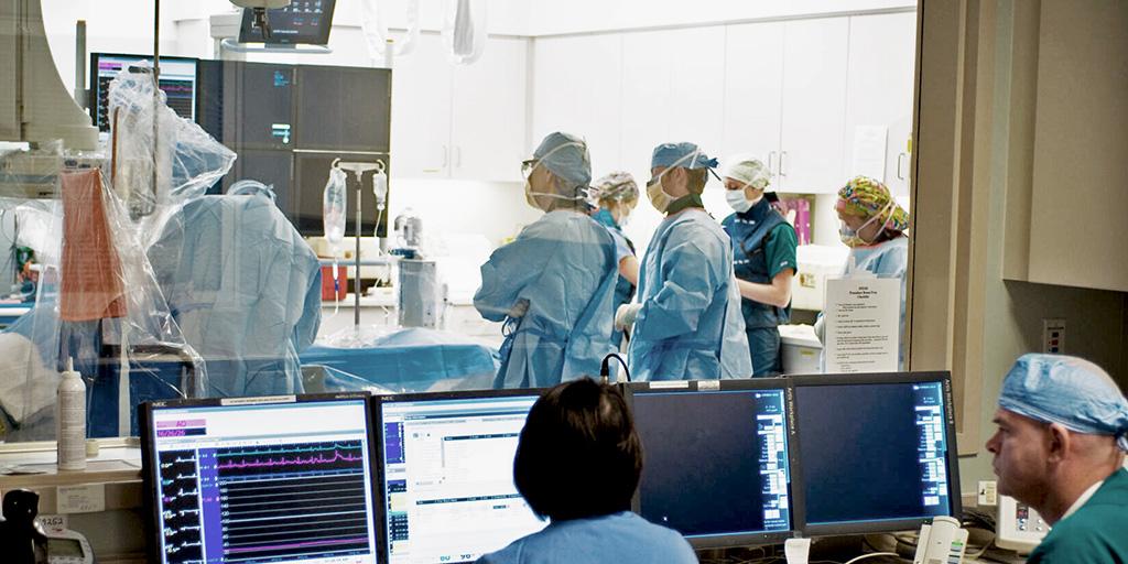 In a cath lab, two people watched on as a group of people surrounded one person while they performed a procedure on a patient.