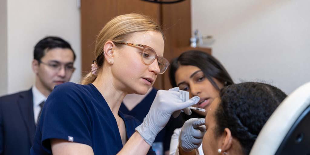 Saranya Wyles, MD, PhD, Dermatology consultant, guides a Dermatology resident in a cosmetic injection practice session while others observe.