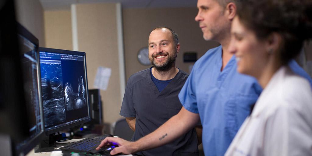 Three medical professionals examine scans on a computer screen.