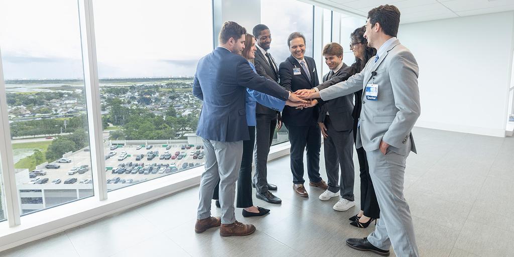 Diagnostic Radiology residents all put their hands together in the center as they stand in a bright hallway area next to a wall of windows.