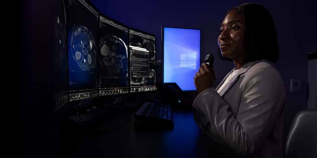 A diagnostic radiologist reviews medical imaging on a computer monitor, while holding up a handheld dictation device.
