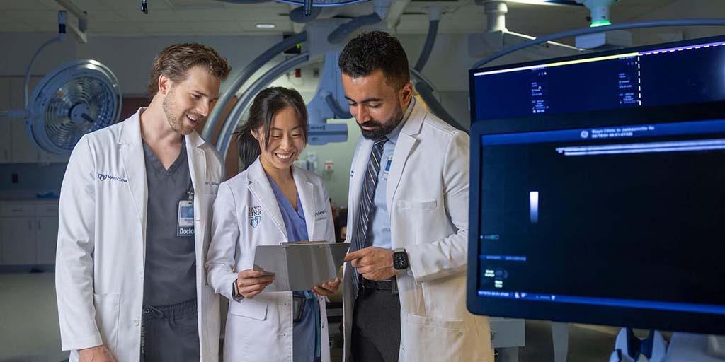 Three radiology residents, wearing white coats, look at a clipboard together while standing in a procedure room.