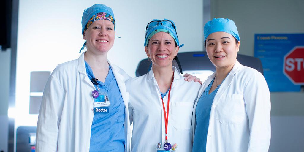 Kendall Tasche, MD; Katie Van Able, MD, and Linda Yin, MD, head and neck surgeons, pose in the operating room prior to Ear, Nose, and Throat (ENT) robotic surgery.