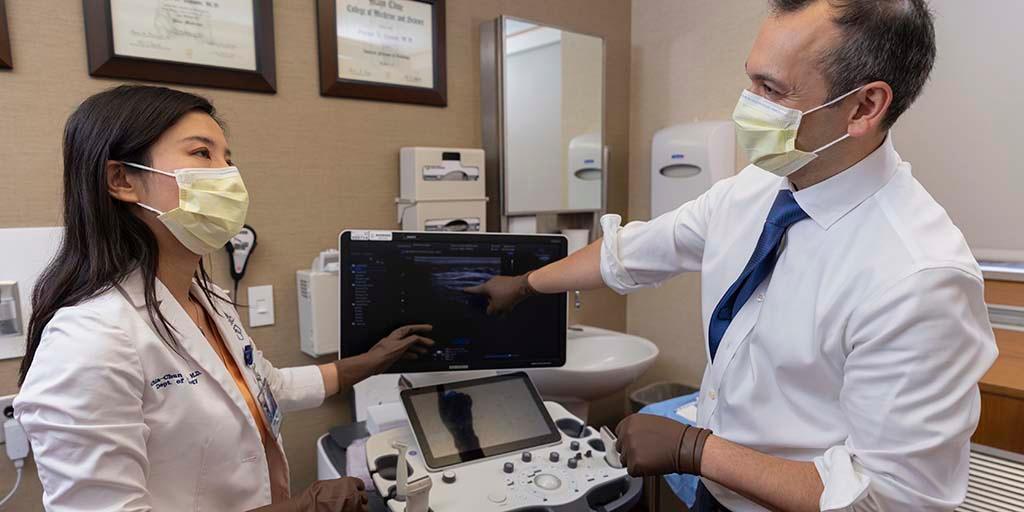 Two neurology consultants, wearing gloves and surgical masks, stand and talk together next to an ultrasound machine, with imaging on the screen.