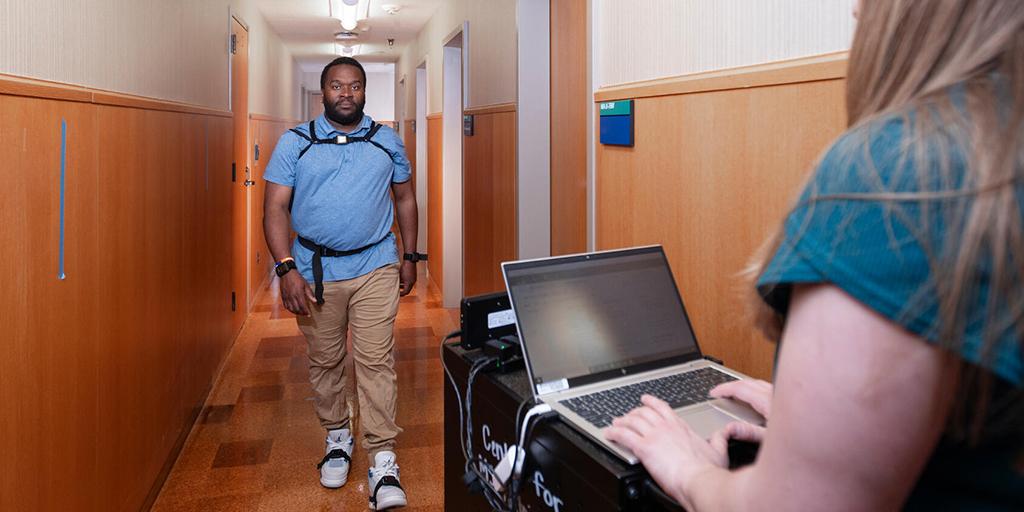 An associate clinical research coordinator, wearing sensors on his wrists, feet, and torso, demonstrates walking down a hallway for gait testing and analysis, for multiple sclerosis (MS) evaluation.