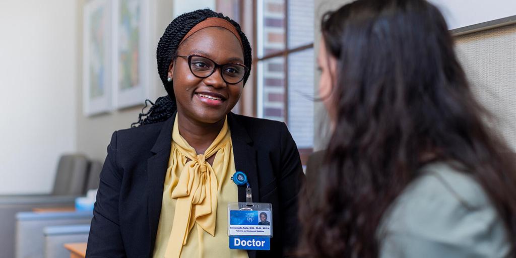 A doctor sits at a desk and talks with an adolescent patient in a doctor's office.