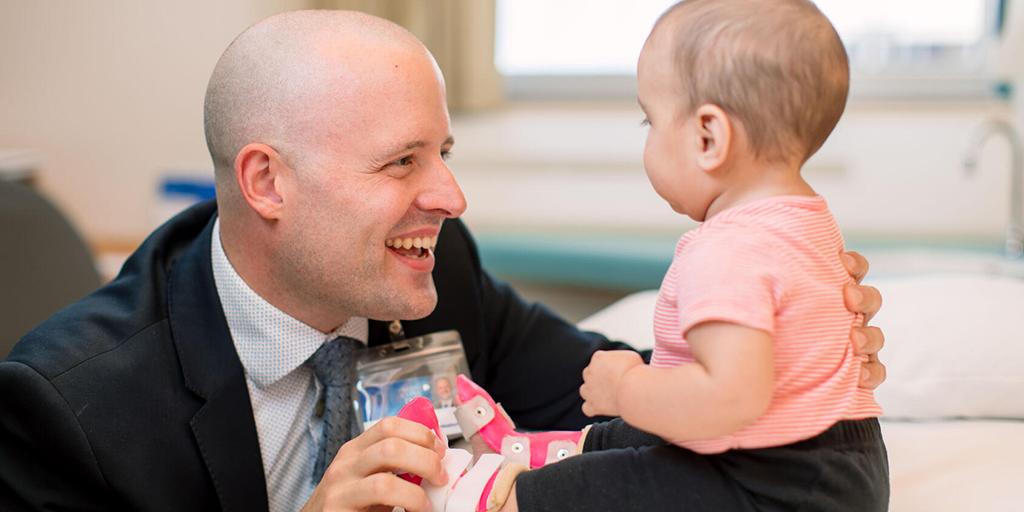 A pediatrician smiles at a baby sitting on an exam table.