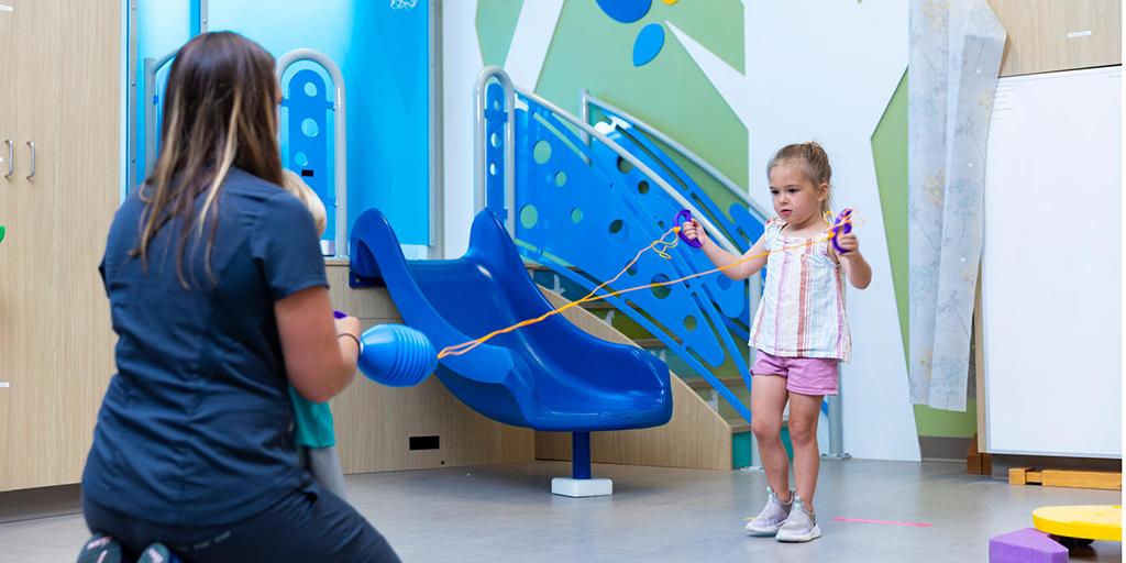 A physical therapist and her young patient use a ball and rope toy in the pediatric gym.