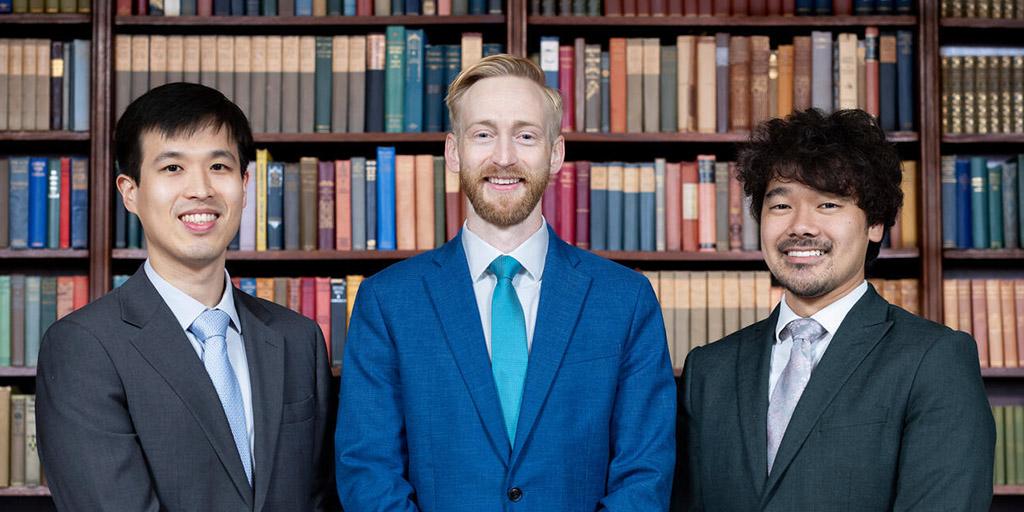 Three 2026 graduates of the program pose for a photo in front of a bookshelf.