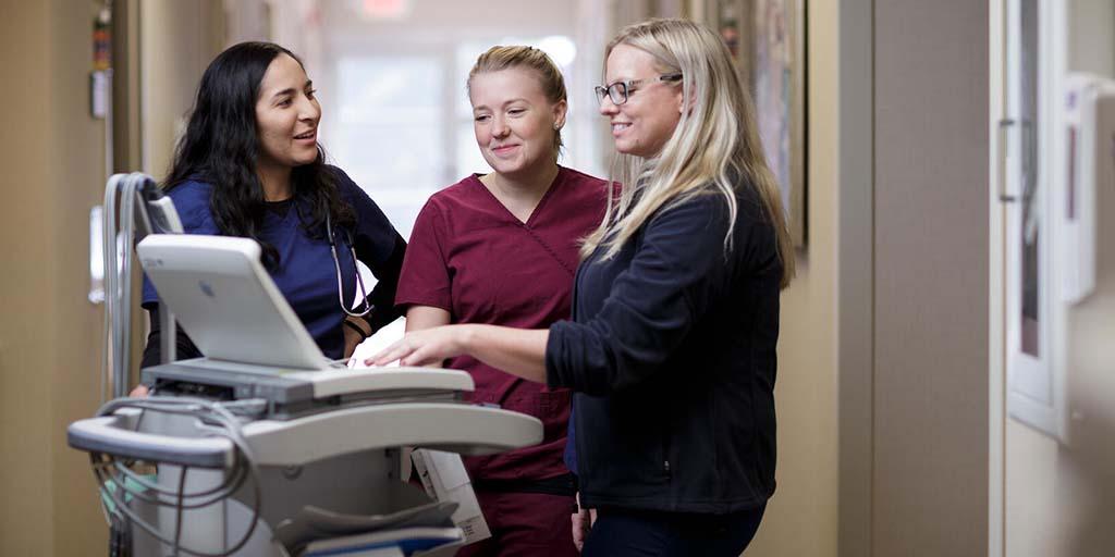 Three health care professionals in a hallway huddled around a standing computer station