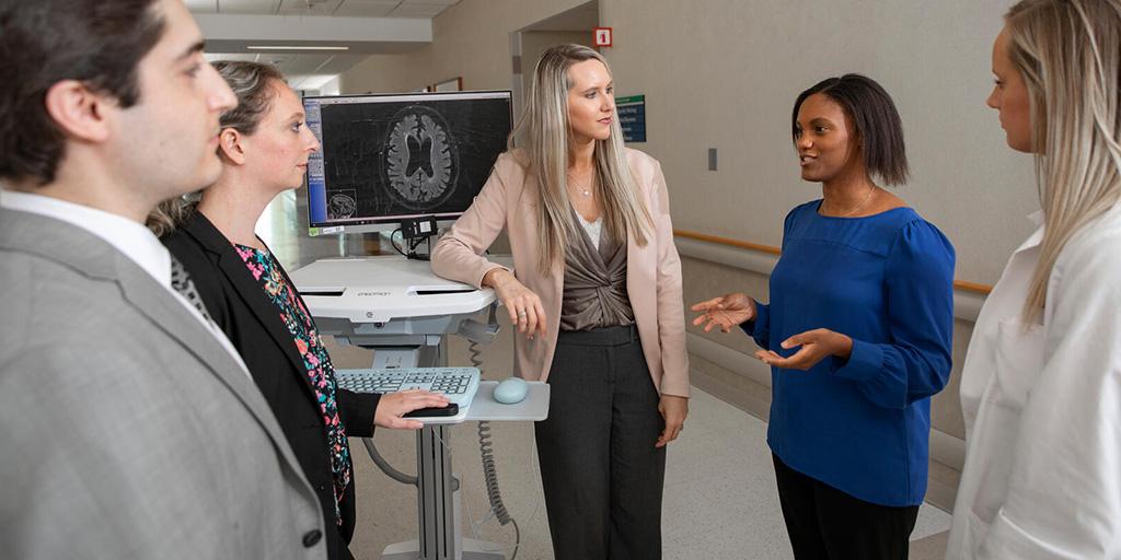 Group of trainees standing in a hallway around a standing workstation discussing something that's on the computer.