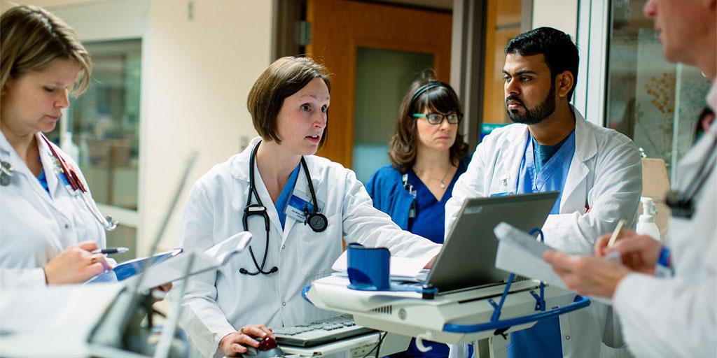 Group of people standing in a hospital hallway with mobile computer stations reviewing what's on the computer and having a discussion