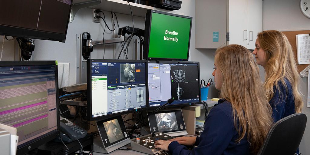 Two students sitting in front of computers in an observation room, providing radiation therapy during simulation lab. 