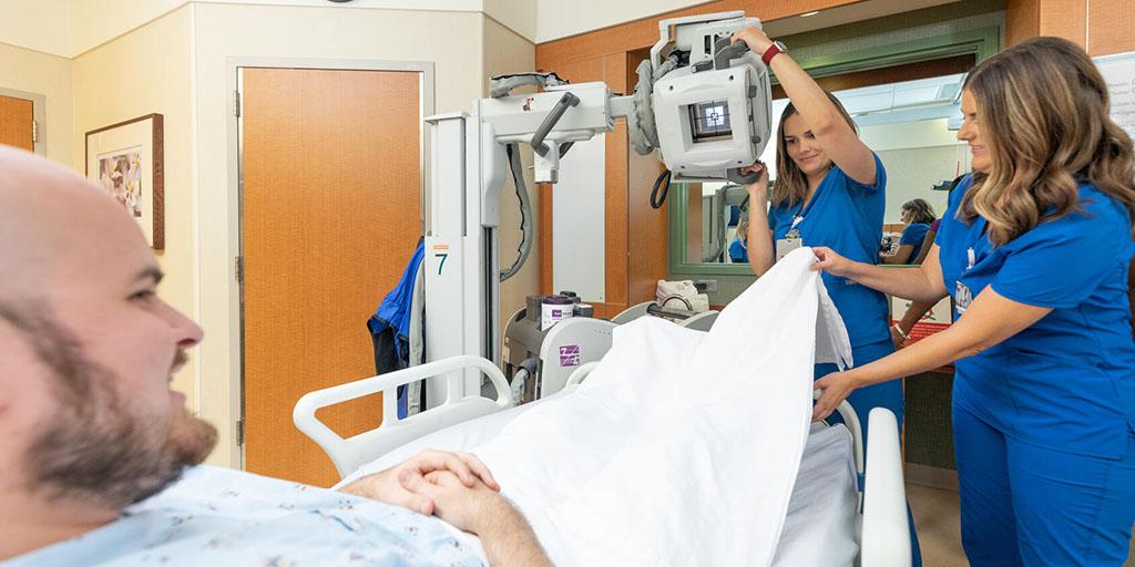 Radiography students practice an X-ray procedure on a practice patient who is in a hospital bed.