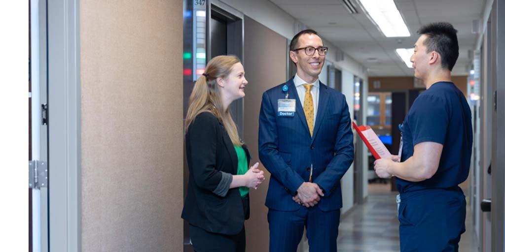 Frank Jing, MD, Dermatology resident, speaks with physicians in a patient hallway. He holds a patient's information sheet in his hand.