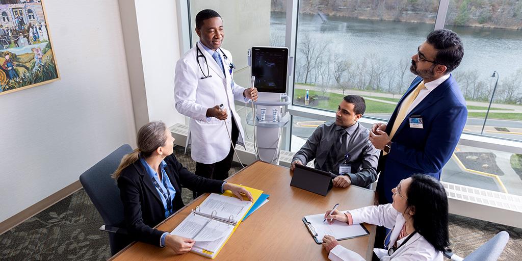 Two people are standing and three people are seated in a conference room having a discussion and looking at a point of care ultrasound machine.