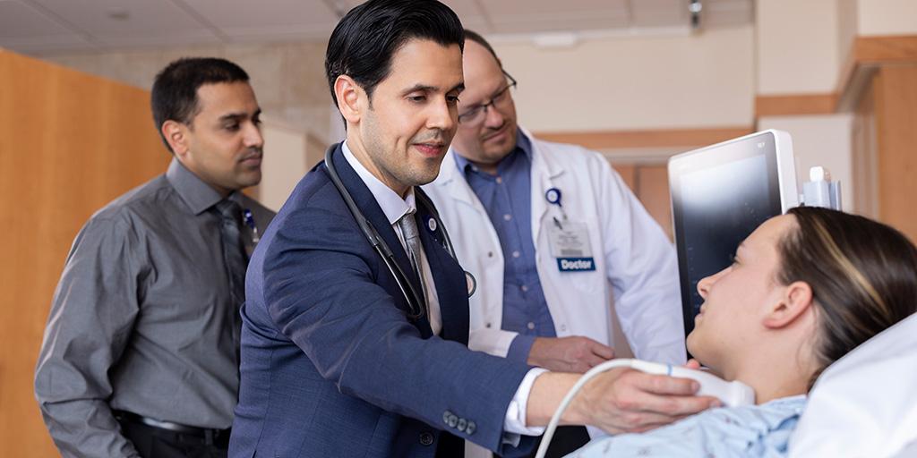 Two doctors stand in the background to assist while one doctor performs an ultrasound procedure on a patient.