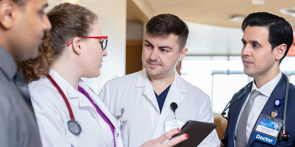 Four doctors stand in the hallway having a discussion while looking at a digital tablet.
