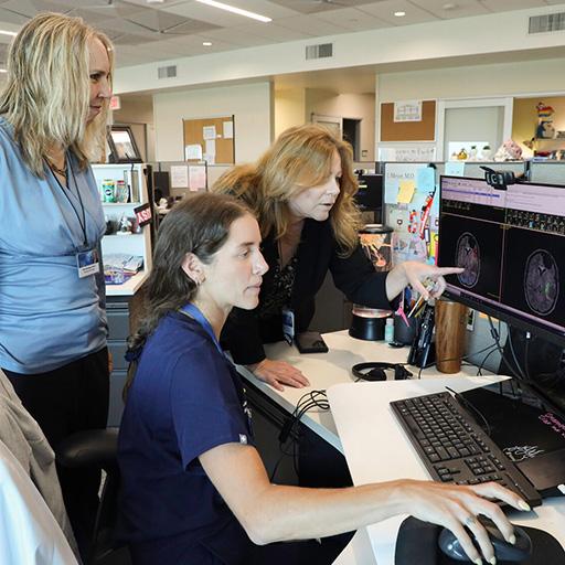 A person sat a desk looking at a computer monitor with two people standing behind them.