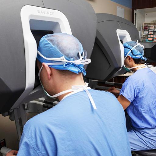 Two surgeons sat side by side, each operating a surgeon's console for a robotic surgical system.