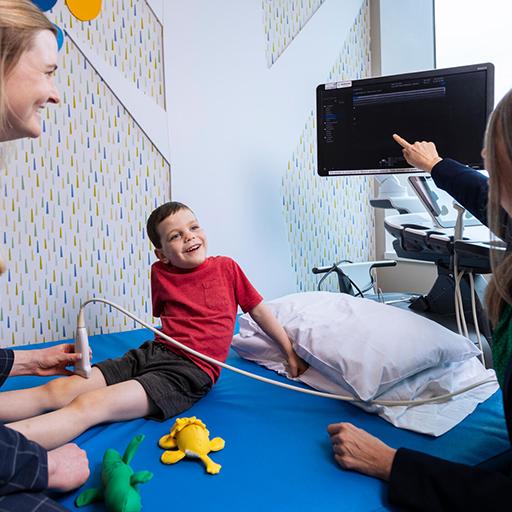 A pediatric rehabilitation fellow and consultant work together with a young patient in a pediatric care room, and do an ultrasound of his knee.