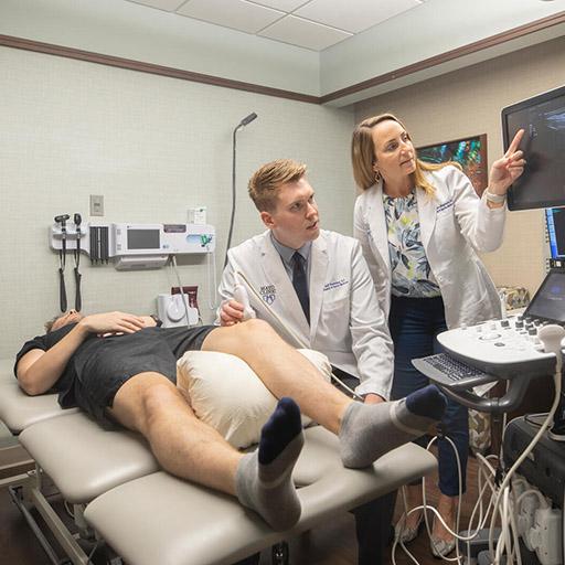 A Sports Medicine resident and a Family Medicine physician perform and ultrasound on a patient's knee in an exam room.