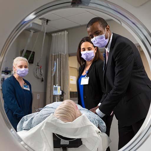 Amir Mbonde, MB, ChB, Resident in Neurology, and Cumara O’Carroll, MD, MPH, Consultant in Neurology, speak with a patient laying on a bed prior to having a brain scan.  Vanesa K. Vanderhye, MSN, CCRN-K, SCRN, Stroke Coordinator in Neurology stands nearby, and all wear masks. This image is taken looking through the scanner with everyone framed in its opening.