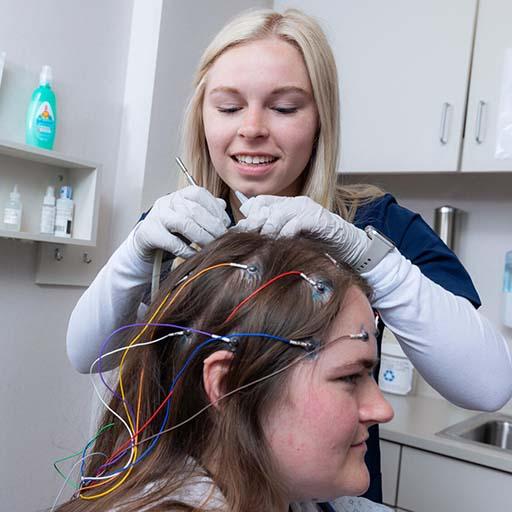A technician attaches head electrodes to a fellow technician's head during an electroencephalogram (EEG) hookup and run study clinical lab session for the Clinical Neurophysiology Technology (CNT) program.