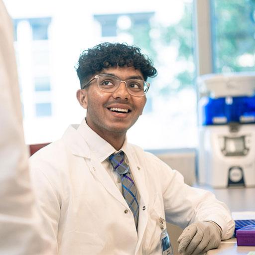 Resident in lab coat in a lab looking up at a colleague and smiling
