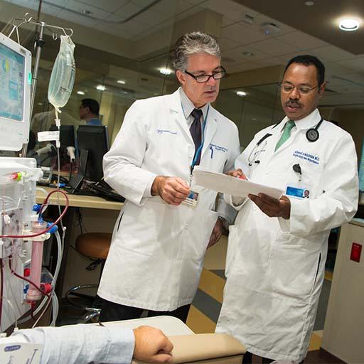Two providers looking at a clipboard, standing near a dialysis machine and patient in a hospital setting
