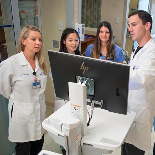 Seven people in white coats standing a hospital hallway examining something on a rolling computer screen.