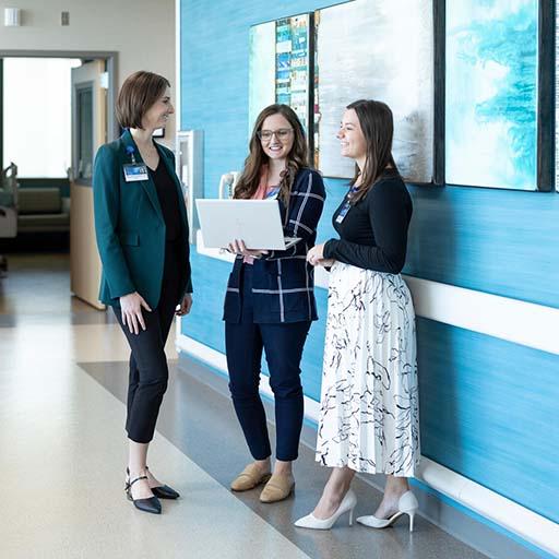 Pharmacists stand with a pharmacy resident and speak in a hallway. One pharmacist holds an open laptop.