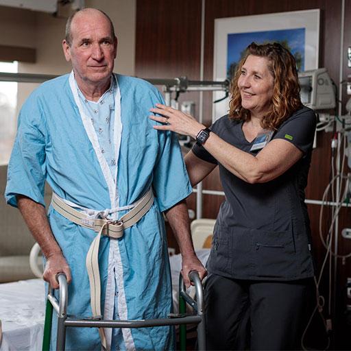 Physical therapist and elderly patient in a hospital room. The PT is assisting the patient to walk with his walker.