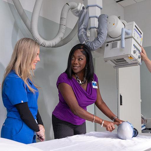 An instructor positions an anatomical model on a table underneath an X-ray machine while radiography students observe.