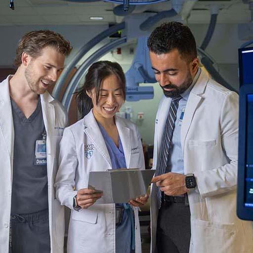 Three radiology residents, wearing white coats, look at a clipboard together while standing in a procedure room.