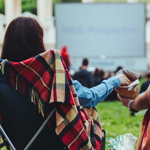 Person sitting in a lawn chair, facing a large outdoor movie screen reaching into a bag of popcorn