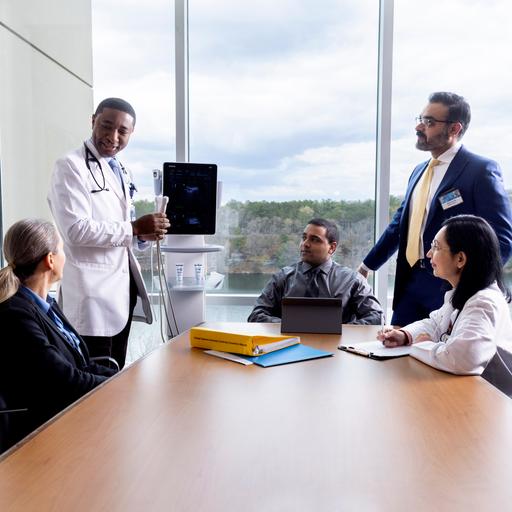 Two people are standing and three people are seated in a conference room having a discussion and looking at a point of care ultrasound machine.