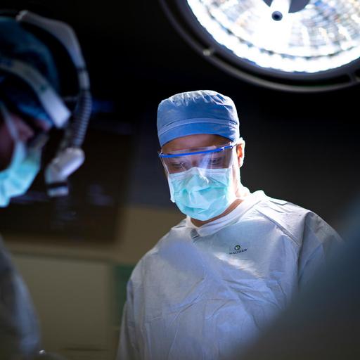 Two doctors in an operating room dressed in medical attire wearing a cap, glasses, and face mask performing a procedure.