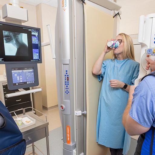 A patient takes a drink while standing in a fluoroscopy imaging machine during a swallowing evaluation.