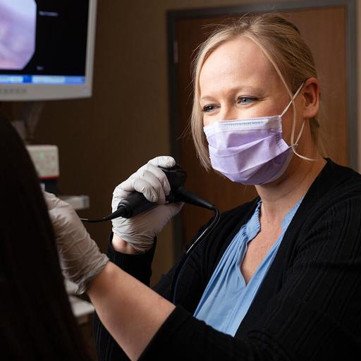 Stacy Holyfield, MS, CCC-SLP, Speech Pathologist, uses a laryngoscope to view a patient's larynx while the image is visible on a screen in the background.