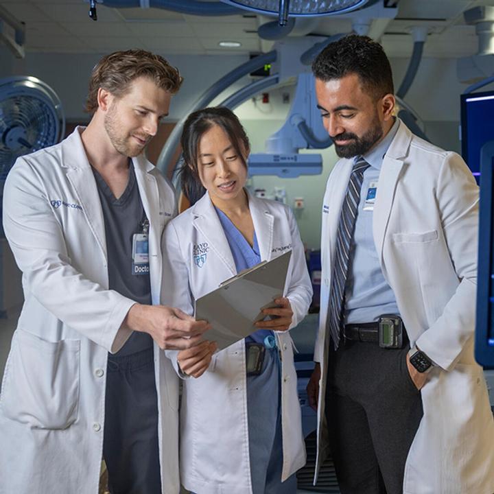 Three radiology residents, wearing white coats, look at a clipboard together while standing in a procedure room.