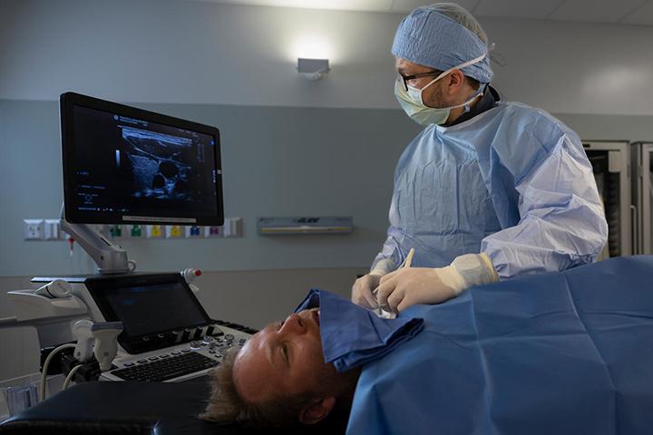 A diagnostic radiologist, wearing scrubs and a surgical mask, simulates an ultrasound exam on a patient's neck.