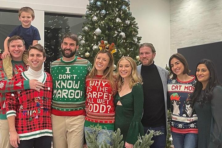 Nine people wearing winter holiday sweaters stood in front of a holiday-decorated tree for a group photo.