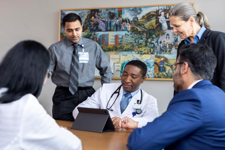 Two people are standing and three people are seated in a conference room having a discussion and looking at a digital tablet.